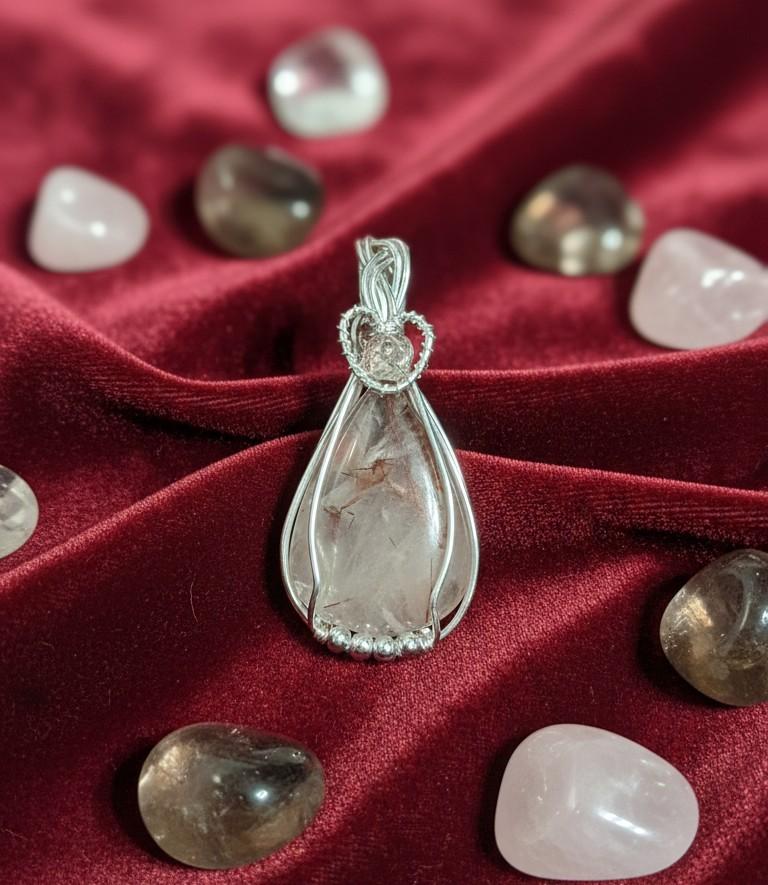 Handgemaakte druppelvormige Rutielkwarts hanger met zilveren wire wrap en een bergkristal kraaltje, gepresenteerd op een luxe rode fluwelen ondergrond.