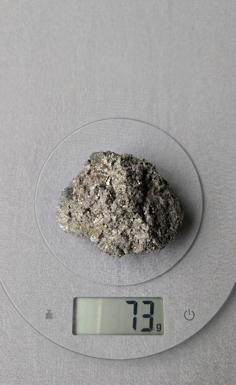 Een compact, ruw cluster van goudkleurige Pyrietkristallen en heldere kwartsdeeltjes, gewogen op een digitale weegschaal die 73 gram aangeeft.