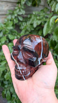 Kristallen schedel van 672 gram in sardonyx agaat met dieprode en bruine aardlagen en een witte band, vastgehouden in de hand.
