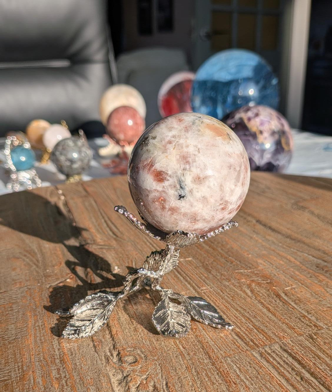 Een gepolijste Zonnesteen bol met warme koperkleurige vlekken, rustend op een gedetailleerde zilverkleurige houder in de vorm van een bloem met bladeren op een houten tafel.