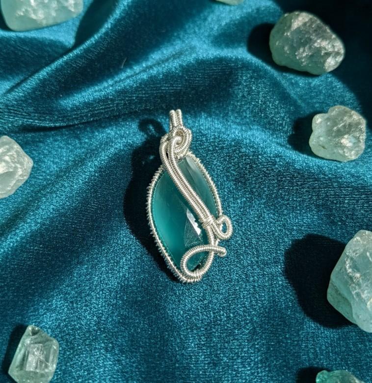 Handgemaakte druppelvormige Serpentijn hanger met een zeegroene kleur en verfijnd zilverkleurig wire wrap vlechtwerk op een blauwe satijnen ondergrond.