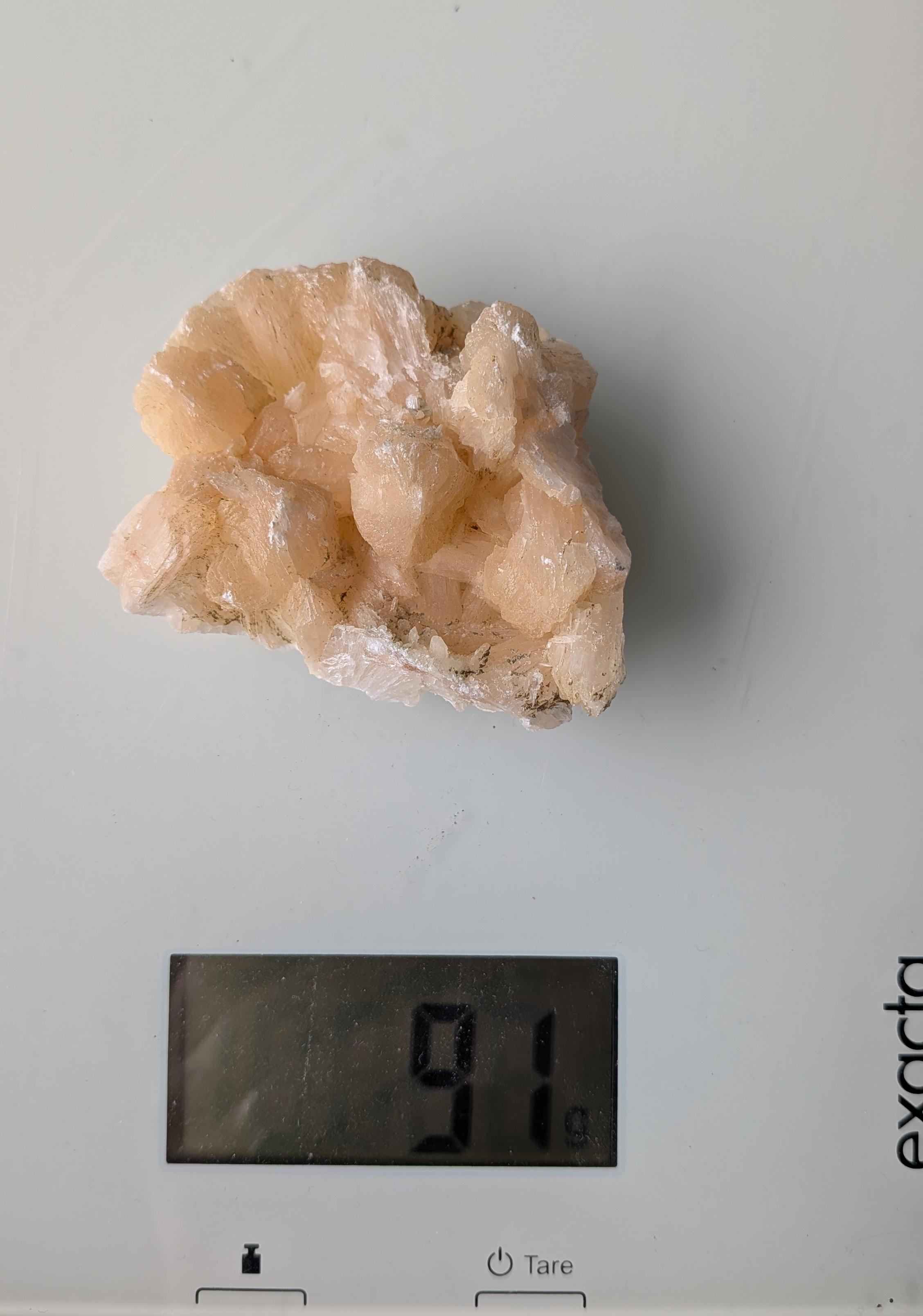 Een natuurlijk, perzikkleurig Stilbiet cluster met waaiervormige kristalstructuren uit Nasik, India, gewogen op een digitale weegschaal die 91 gram aangeeft.