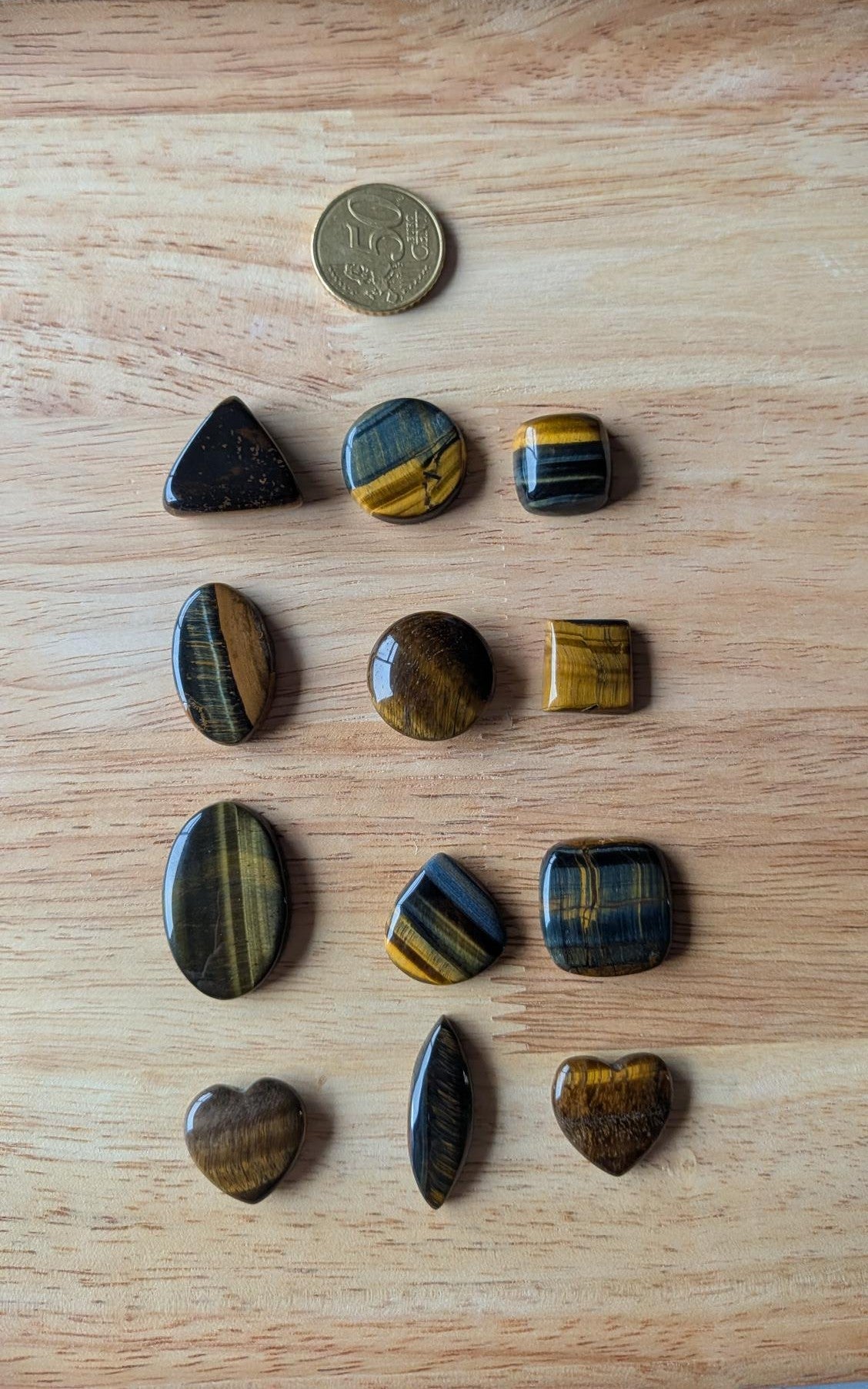 Twaalf verschillende vormen Tijgeroog edelstenen, waaronder hartjes, driehoeken en ovalen, op een houten achtergrond naast een 50 cent muntstuk.