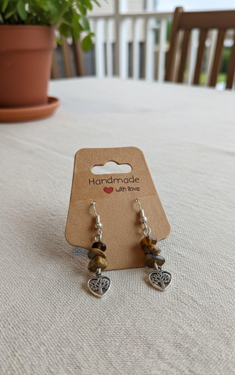 Handgemaakte zilverkleurige oorbellen met een levensboom in een hartje en bruine tijgeroog splitkralen op een kraft kaartje.