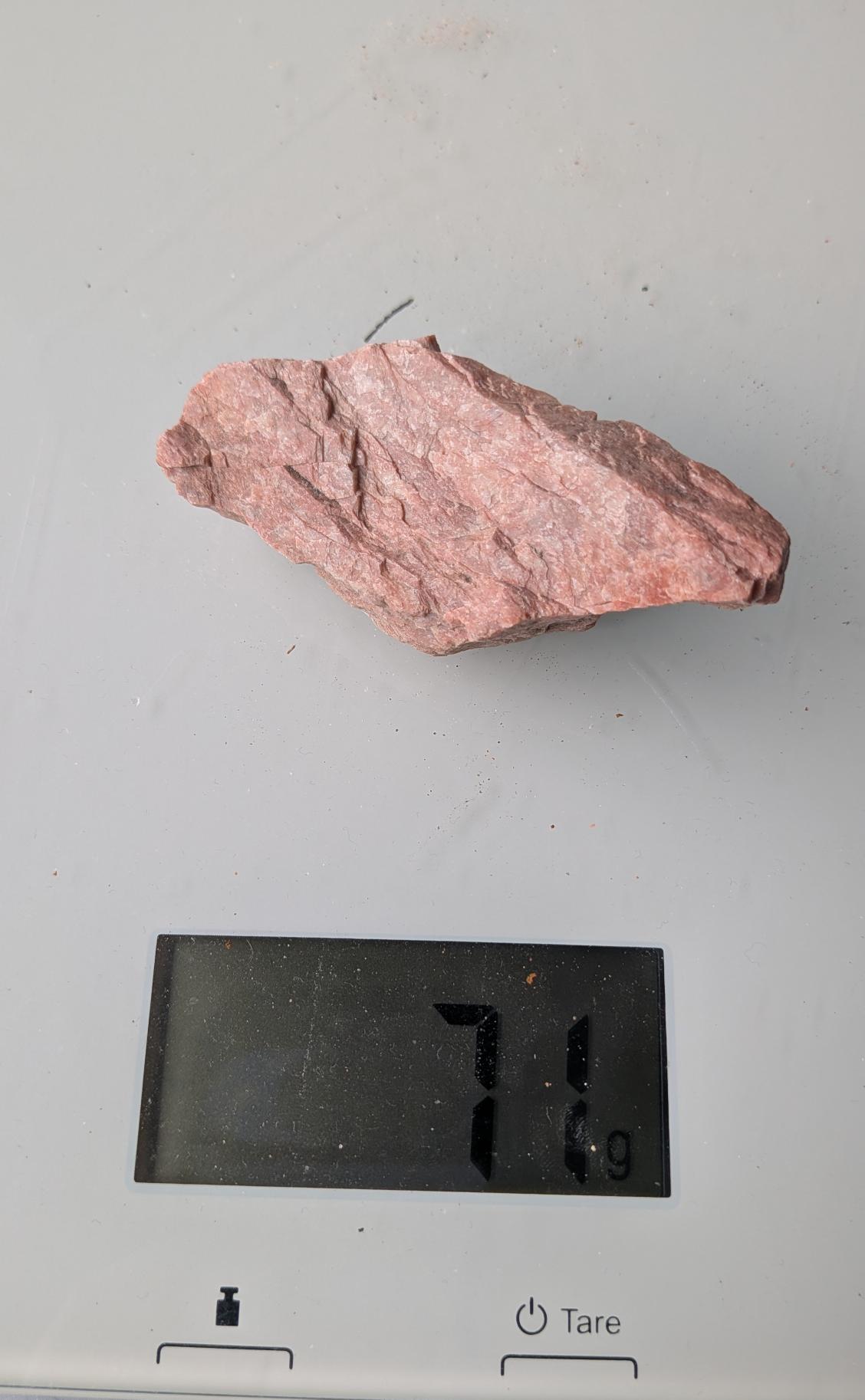 Een ruw stuk rode Veldspaat van 71 gram met een karakteristieke, licht glinsterende structuur, gefotografeerd op een digitale weegschaal.