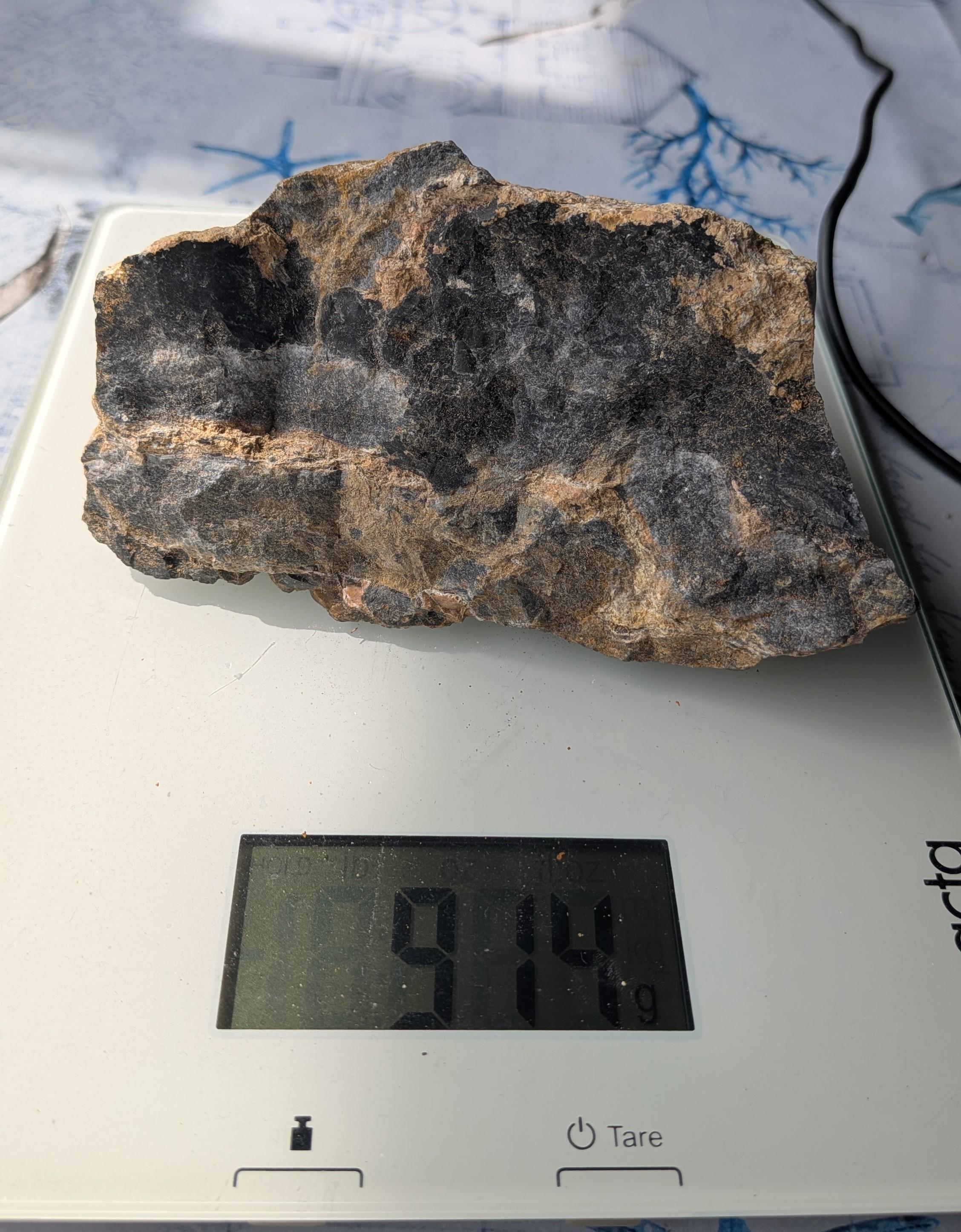 Een massief stuk Vuursteen in matrix van 914 gram met een zwarte, glanzende kern en een ruwe, bruine buitenkant, gefotografeerd op een digitale weegschaal.