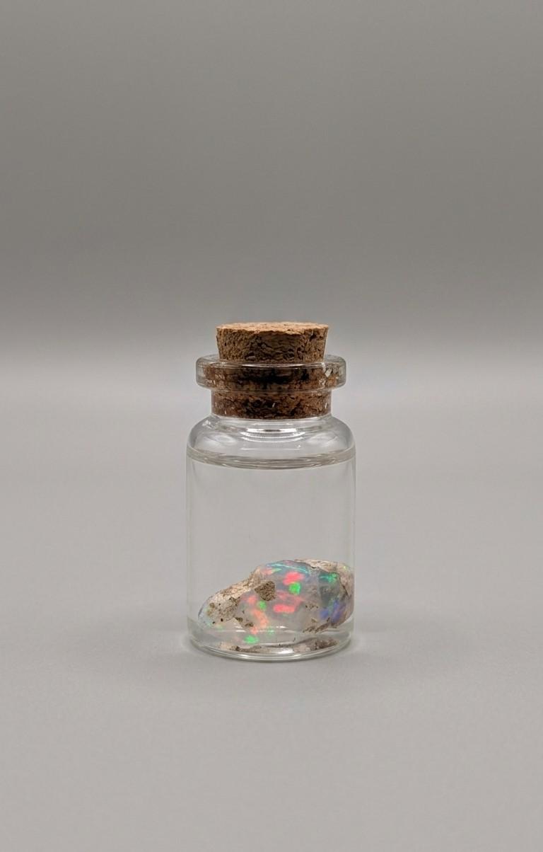 Uitzonderlijke Ethiopische Water Opaal met intens confetti-achtig kleurenspel van rood, groen en blauw, gepresenteerd in een 37 mm glazen display-potje met water.