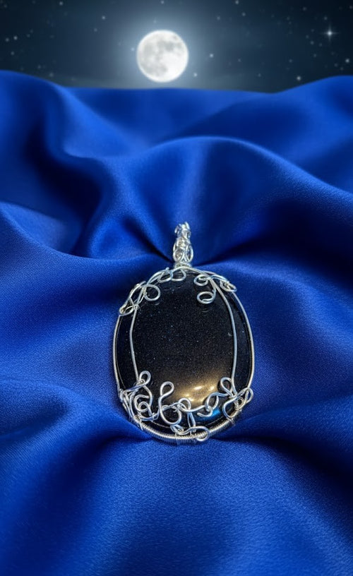 Een zeer grote ovale hanger van fonkelende zwarte goudsteen met een gedetailleerde zilveren wire-wrap wikkeling, rustend op golvende blauwe stof onder een volle maan.