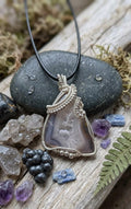 Handgemaakte bruin-witte Agaat hanger met zichtbare kristallen en verzilverd wire wrap vlechtwerk met een spiraal, gepresenteerd op donkere stenen.