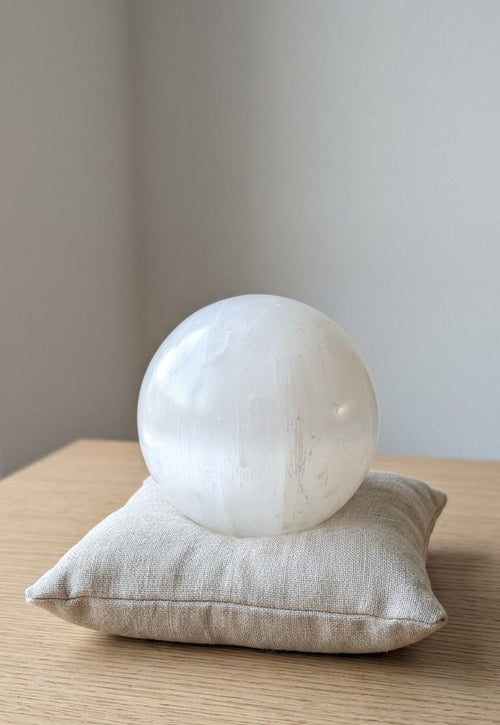 Grote satijnwitte Seleniet bol van 7,5 cm op een beige kussentje. Kristal voor zuivering, rust en spirituele reiniging.