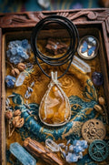 Druppelvormige geel-bruine Agaat hanger met gedetailleerd verzilverd wire wrap werk, gepresenteerd in een houten kistje met steampunk elementen, lavendel en kristallen.