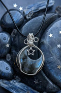 Handgemaakte maanvormige Labradoriet hanger met blauwe glans, verzilverd wire wrap vlechtwerk en een klein sterretje, gepresenteerd op donkere stenen met zilveren ster-accenten.
