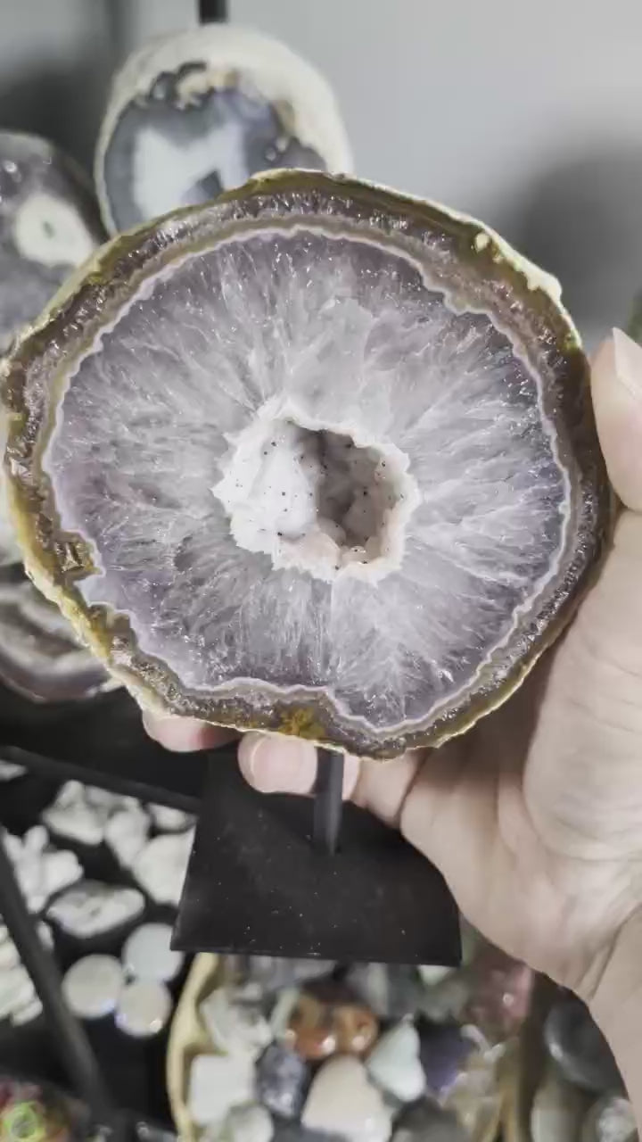 Een hand houdt een grote Agaat-geode van 1237 gram op een zwarte standaard vast, met zicht op het kristalhar