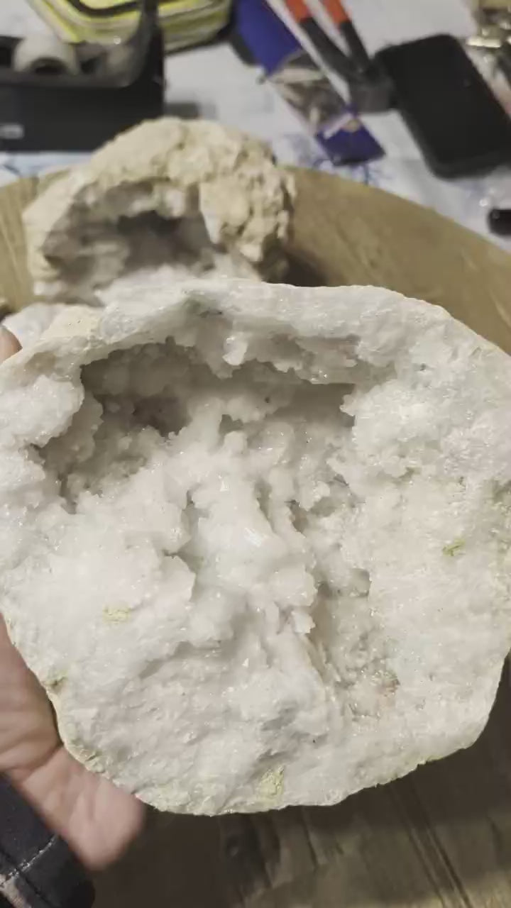 Brede, platte witte kristalgeode van 1155 gram met een groot oppervlak aan schitterende kristallen, liggend op een houten ondergrond.