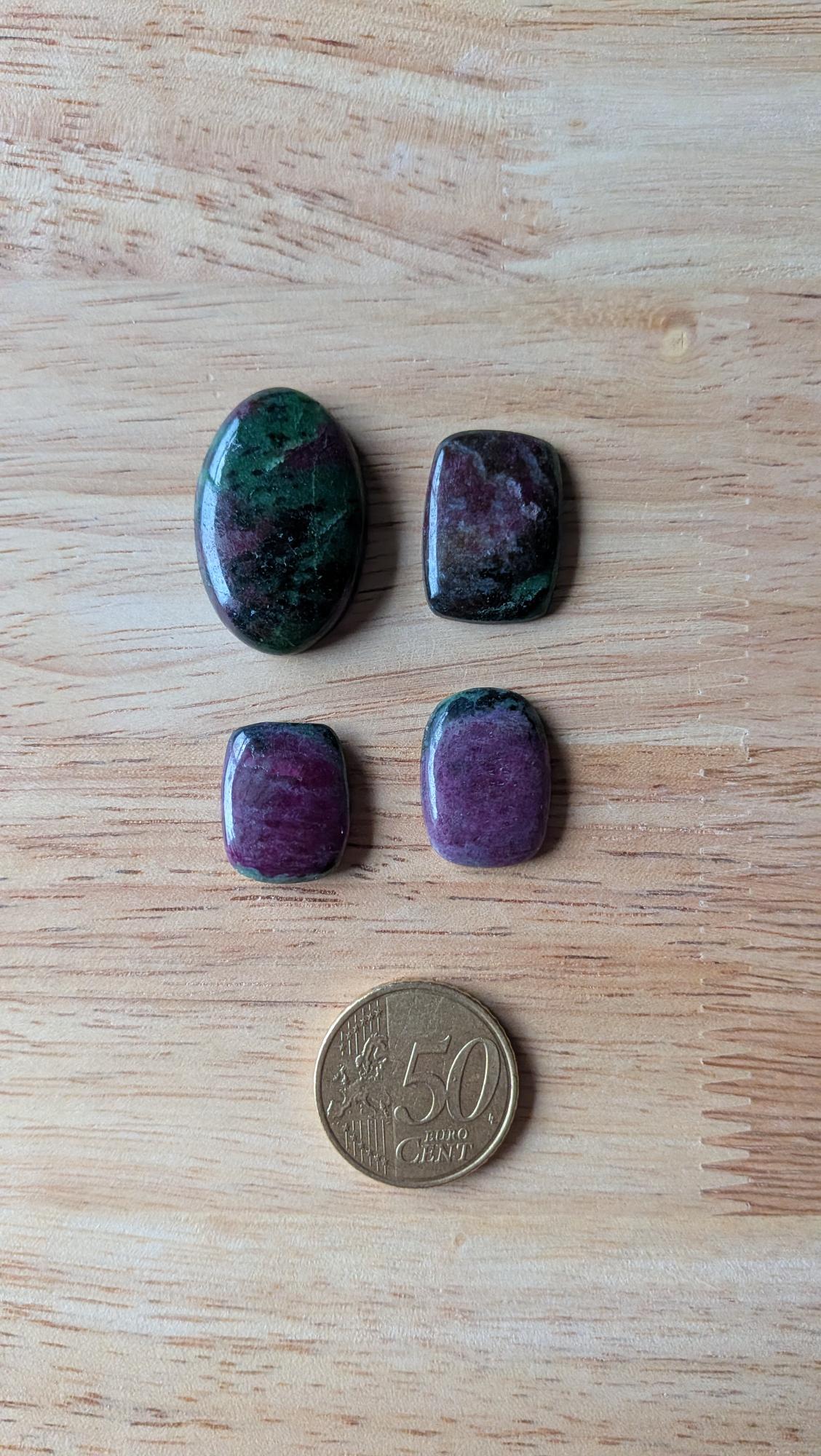 Vier gepolijste robijn in zoisiet edelstenen in ovale en rechthoekige vormen met rood-groene patronen naast een 50 eurocent munt op een houten ondergrond.