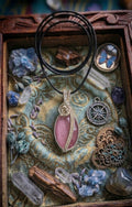 Handgemaakte facetgeslepen Rozenkwarts hanger met een verzilverd wire wrap roosje, liggend in een decoratief houten kistje met blauwe vlinder en kristallen.