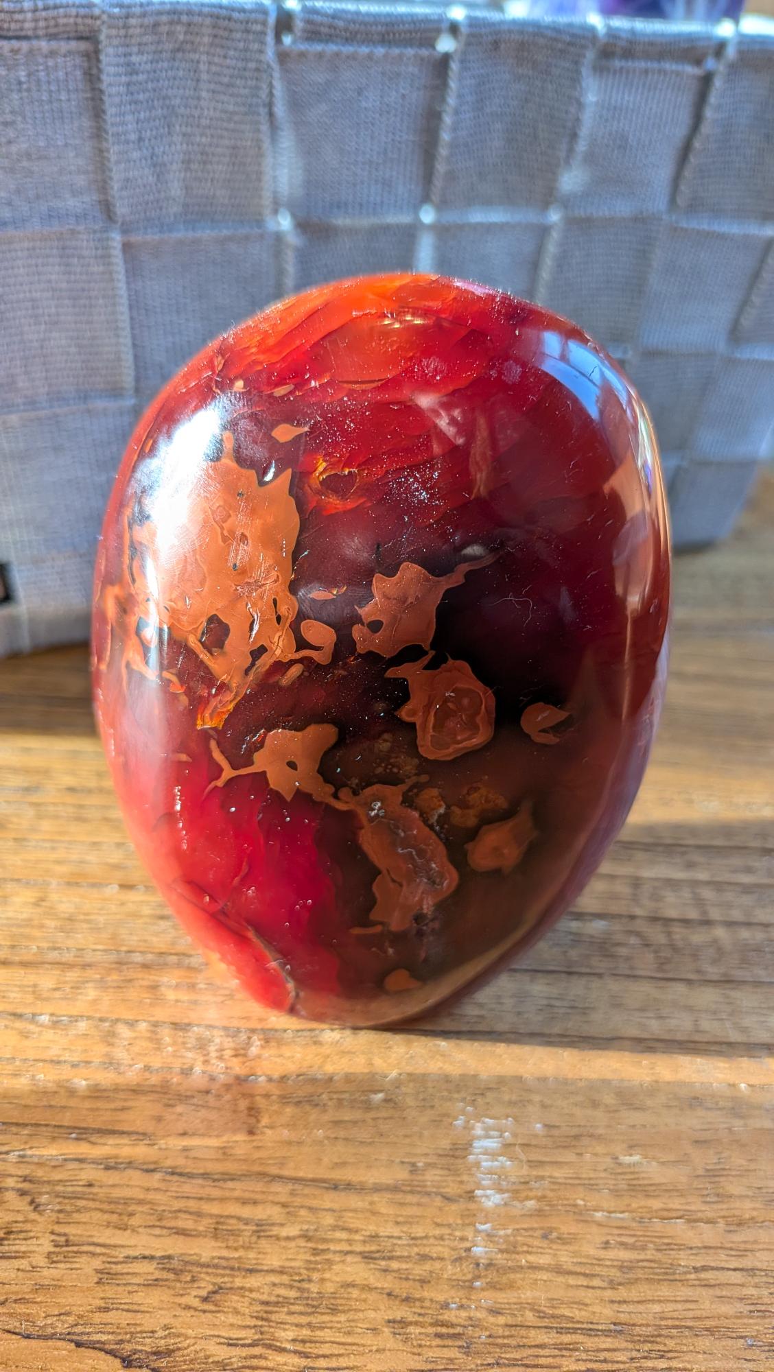 "Gepolijste XL Carnéool vrije vorm edelsteen van 580 gram met diepe rode en vurige oranje kleurschakeringen, gefotografeerd op een natuurlijke houten ondergrond."