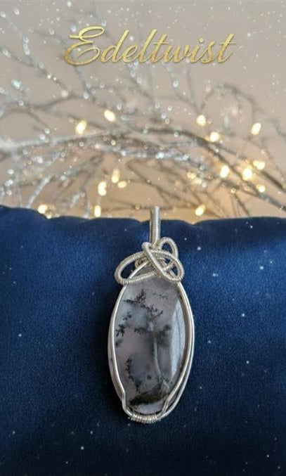 Handgemaakte zilverkleurige wire-wrapped hanger van Edeltwist met een ovale Dendriet Agaat, tonend natuurlijke zwarte boomachtige insluitingen op een grijze basis, gepresenteerd op een blauw fluwelen kussen.