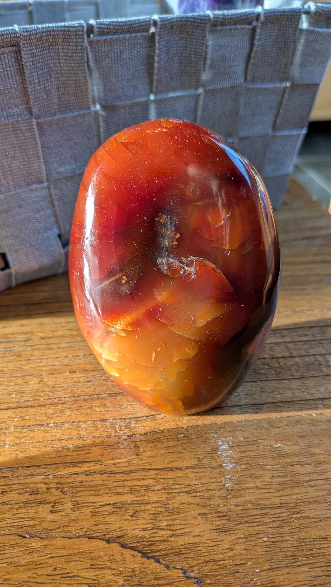 "Gepolijste XL Carnéool vrije vorm edelsteen van 580 gram met diepe rode en vurige oranje kleurschakeringen, gefotografeerd op een natuurlijke houten ondergrond."