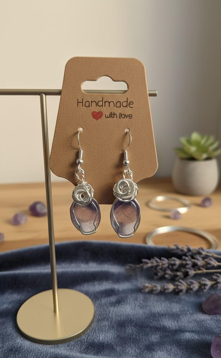 Twee handgemaakte zilverkleurige wire wrapped oorbellen met paarse fluoriet edelstenen hangend aan een bruin kaartje met de tekst 'Handmade with love'.