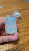"Hand die een Aqua Aura Kwarts punt vasthoudt met een opvallende blauwe glans op een houten tafeloppervlak."