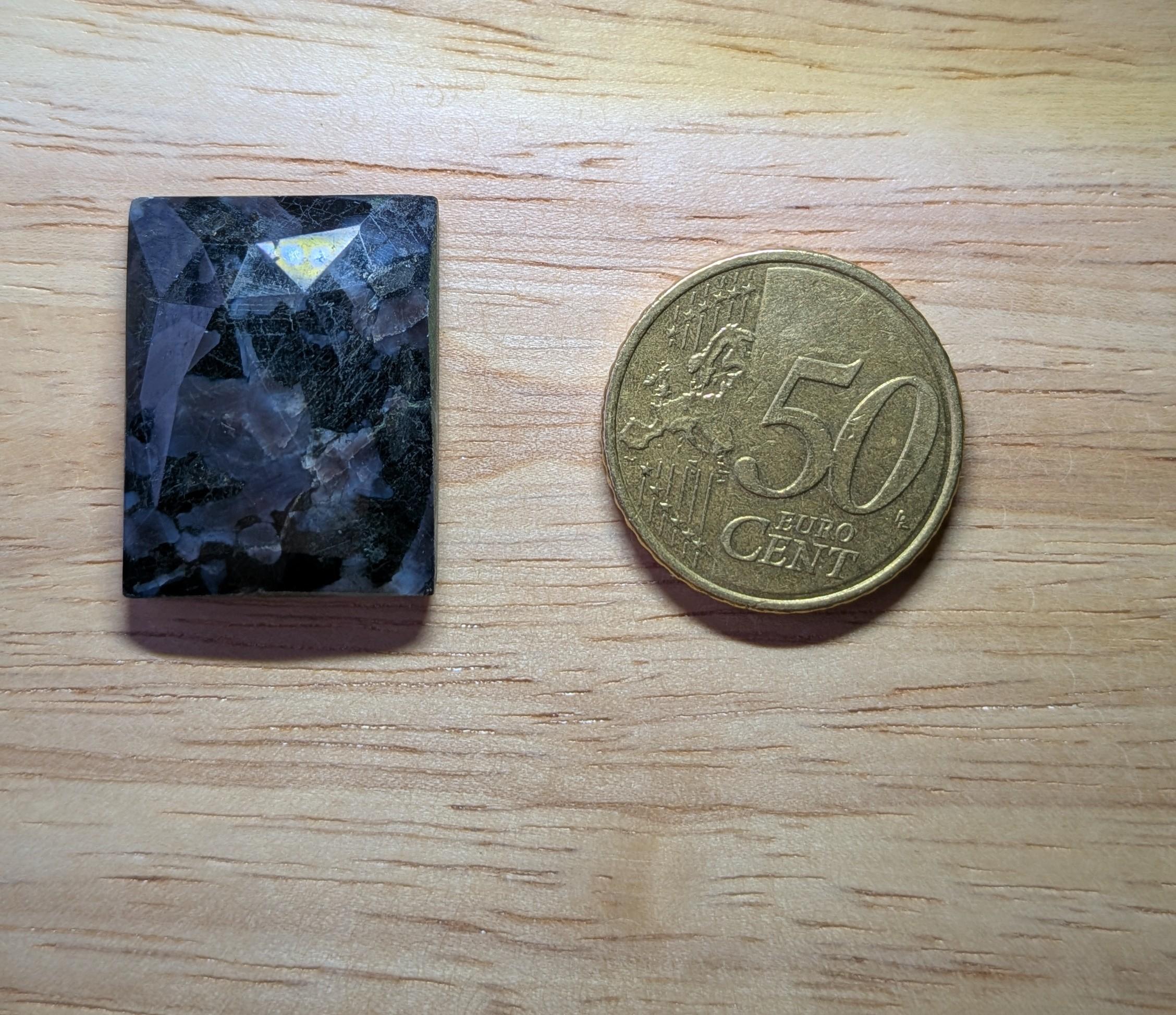 Gefacetteerde rechthoekige Sodaliet edelsteen met donkerblauwe en grijze patronen naast een munt ter schaalvergelijking.