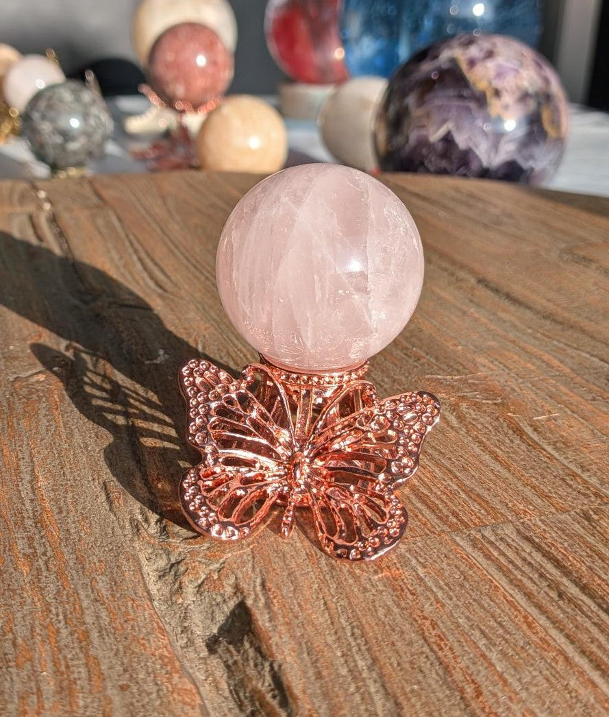 Een zachtroze gepolijste Rozenkwarts bol geplaatst op een glanzende roségoudkleurige houder in de vorm van een vlinder, staand op een rustieke houten tafel in het zonlicht.