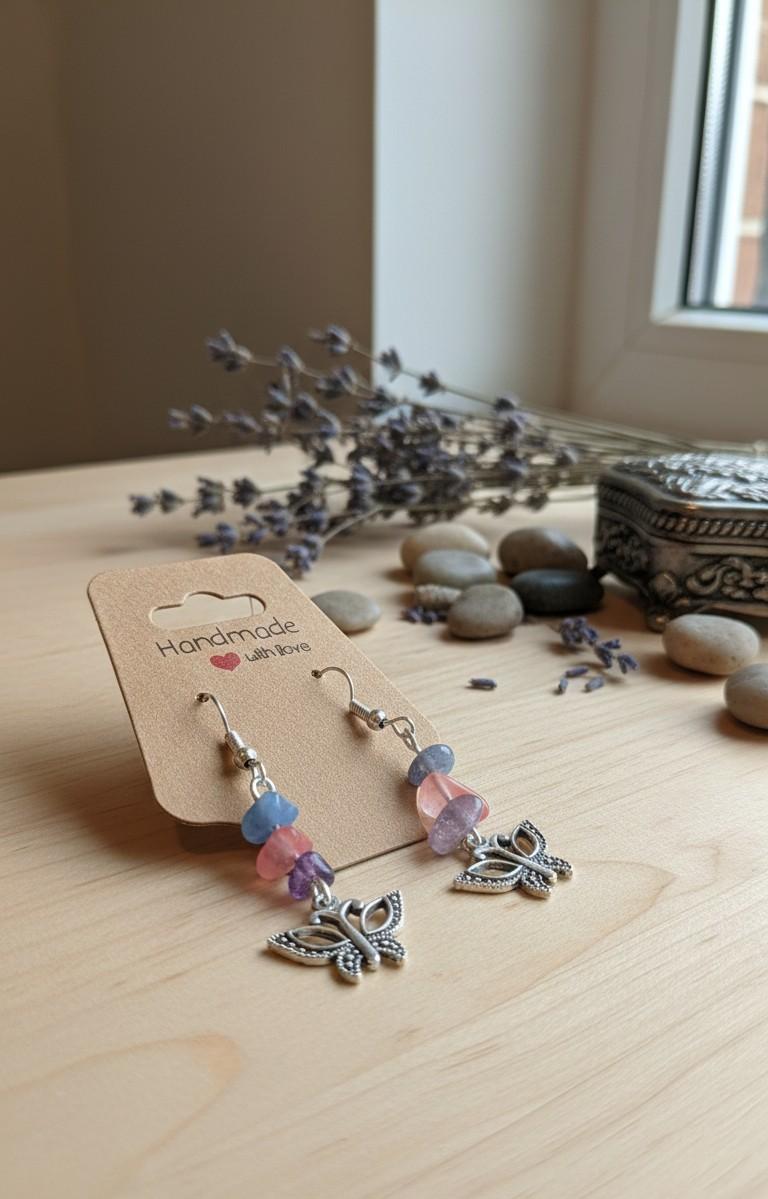 Handgemaakte zilverkleurige oorbellen met vlinderbedeltje en pastelkleurige edelsteen splitkralen op een kraftkarton verpakking.