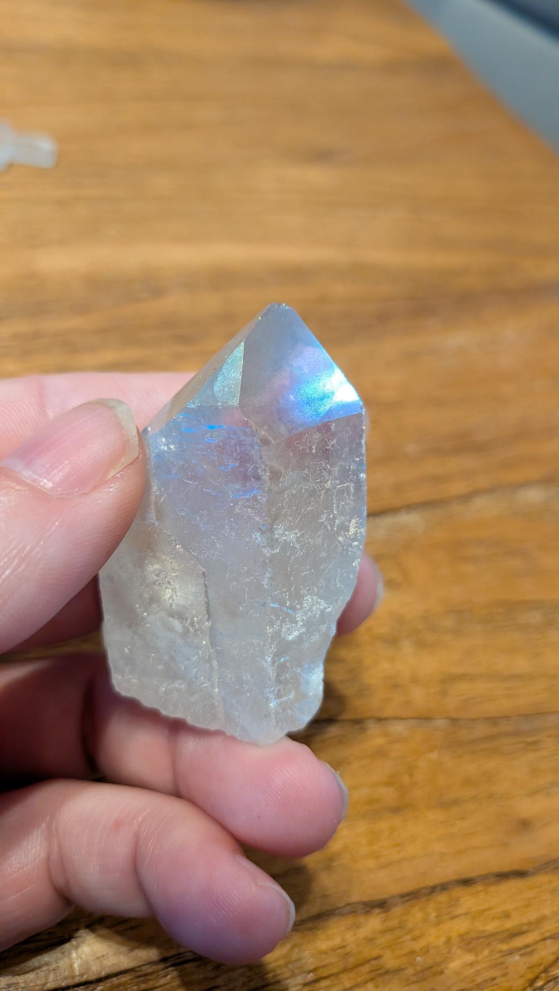 "Close-up van een iriserende Angel Aura Kwarts punt met blauwe en roze regenboogglans vastgehouden in een hand tegen een houten achtergrond."