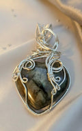 Handgemaakte zilverkleurige wire-wrapped hanger met een hartvormige Labradoriet edelsteen die een mystieke groen-gouden glans toont, gepresenteerd op een lichte zijden ondergrond.