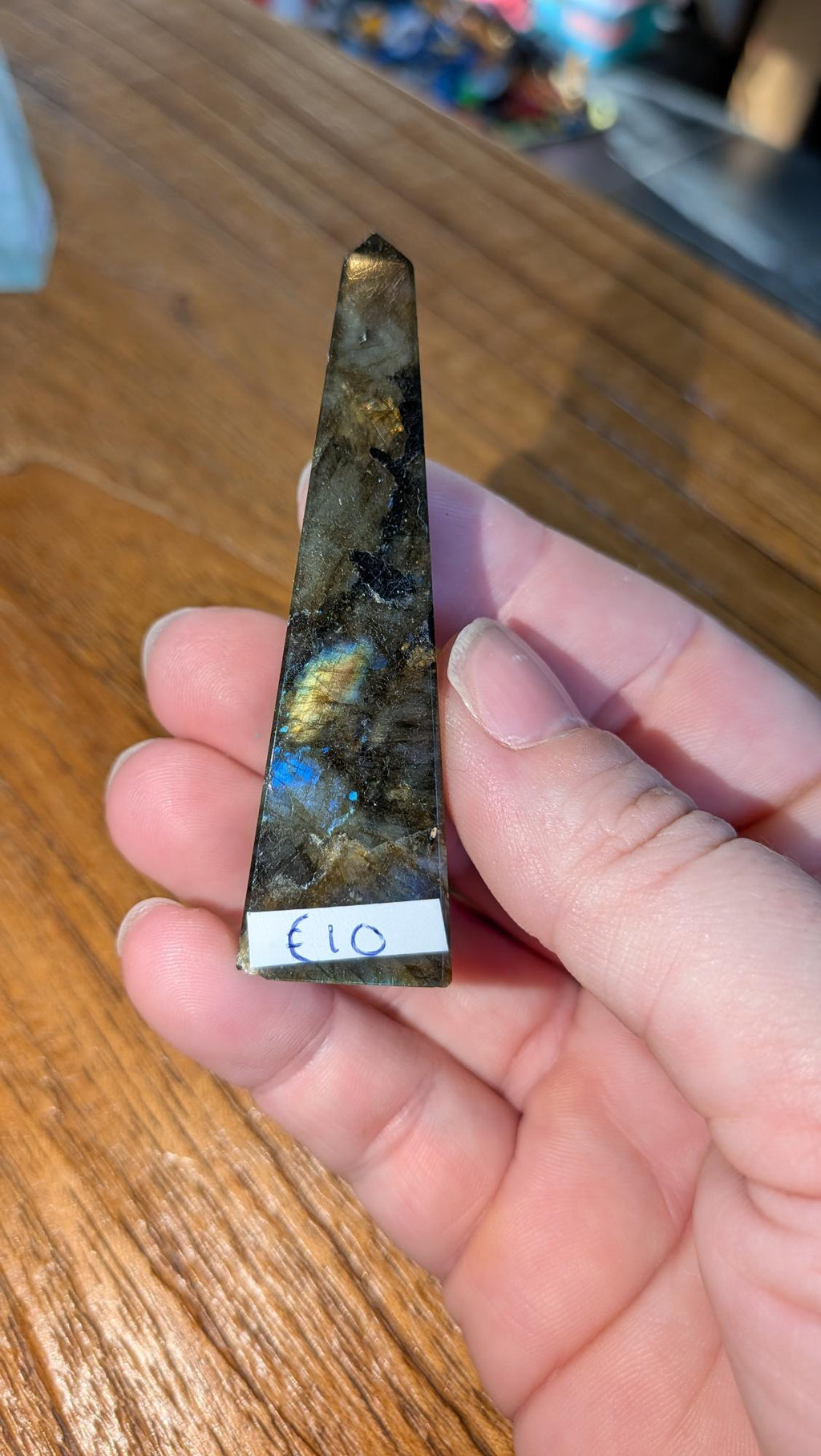 "Slanke gepolijste labradoriet toren van 6,5 cm met een opvallende blauwe en gouden glans, vastgehouden in een hand tegen een houten achtergrond."