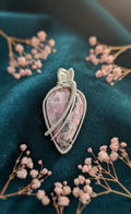 Handgemaakte wire-wrapped hanger van roze Rhodoniet met zilverkleurig draad, liggend op donkergroen fluweel en omringd door kleine roze droogbloemen