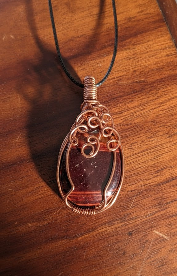 Handgemaakte koperen wire-wrapped hanger met een gepolijste bruin-rode Tijgeroog edelsteen, liggend op een houten ondergrond met artistieke koperen krullen aan de bovenzijde.