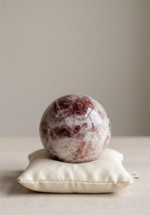 Gepolijste Zonnesteen bol van 5 cm met witte en roodbruine patronen op een wit kussentje, prijs 27,50 euro. Kristal voor zelfvertrouwen en positiviteit.